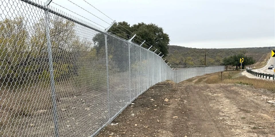 Long galvanized chain link security fence with barbed wire arms along a cleared line in Castroville, Texas